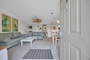 A living room with a sofa, coffee table, and a striped rug. There is a ceiling fan and a dining area with a table and chairs. - Shorehaven Lakefront & Steps to Beach w Pool (North Myrtle Beach)