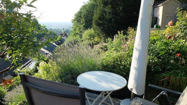 A patio with a table and chairs, surrounded by lush greenery and a view of a town. - Quiet, cozy vacation apartment near the forest. (Neustadt an der Weinstraße)