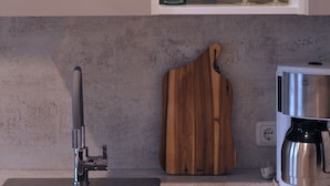 A kitchen with a stainless steel sink, a wooden cutting board, a coffee maker, and shelves with various kitchenware including mugs and wine glasses. - Quiet, cozy vacation apartment near the forest. (Neustadt an der Weinstraße)
