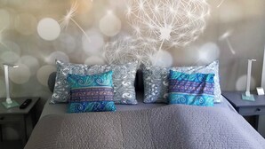 A bed with patterned pillows and a textured blanket, a bedside table with a lamp, and a wall with a dandelion pattern. - Quiet, cozy vacation apartment near the forest. (Neustadt an der Weinstraße)