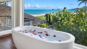 Ein modernes Badezimmer mit einer großen Badewanne, gefüllt mit Rosenblüten, Meerblick und Surfbrettern, die an der Decke befestigt sind.