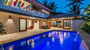 Ein modernes Haus mit Pool, einem Balkon mit Surfbrettern und einem tropischen Garten.