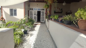 Un espacio al aire libre techado con piso de baldosas, plantas en macetas y una puerta blanca que da a un edificio.