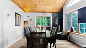 A dining area with a wooden ceiling, a fireplace, and large windows with a view of greenery.