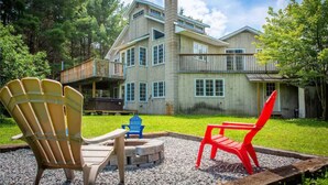 A large, multi-story house with a prominent chimney, a deck, and a fire pit area with two Adirondack chairs.