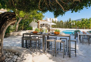 An outdoor dining area with stone flooring, wooden tables, and chairs, surrounded by greenery and a pool.