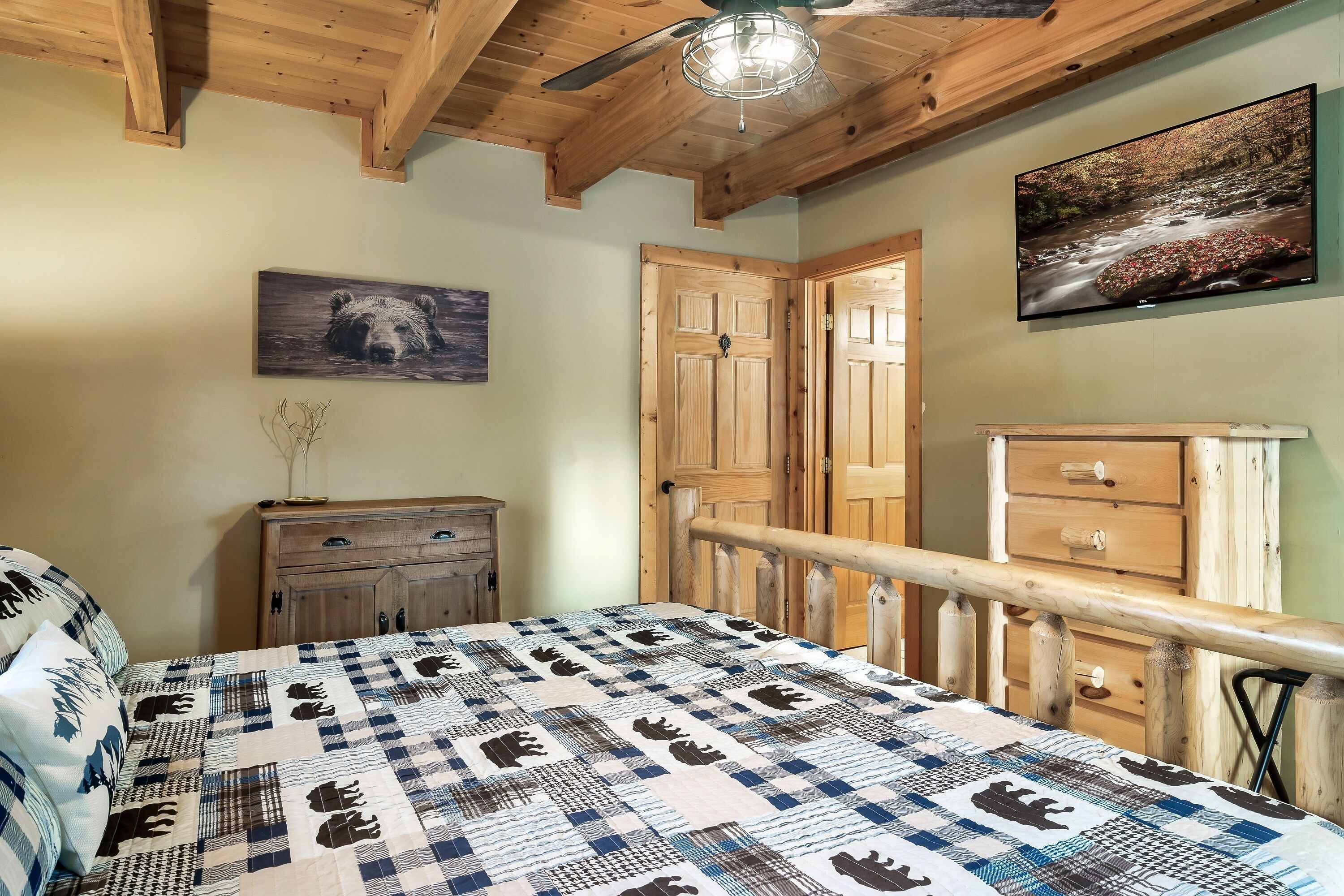 A bedroom with a checkered bedspread, wooden ceiling beams, a ceiling fan, a painting of a bear, a wooden dresser, and a door with a glass panel.