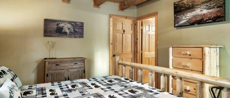 A bedroom with a checkered bedspread, wooden ceiling beams, a ceiling fan, a painting of a bear, a wooden dresser, and a door with a glass panel.