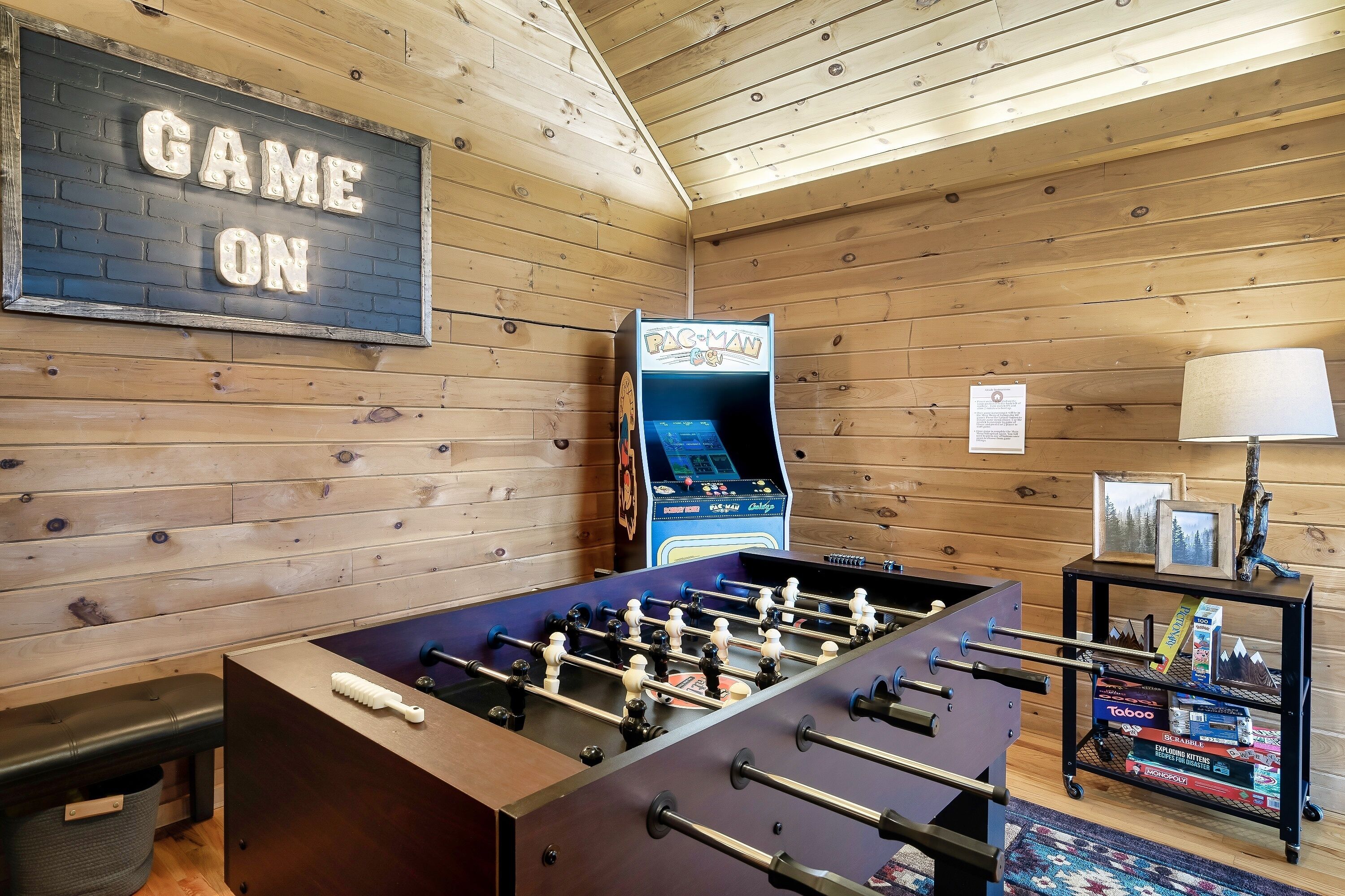 A foosball table, a Pac-Man arcade machine, and a foosball table in a wooden room with a 'GAME ON' sign.