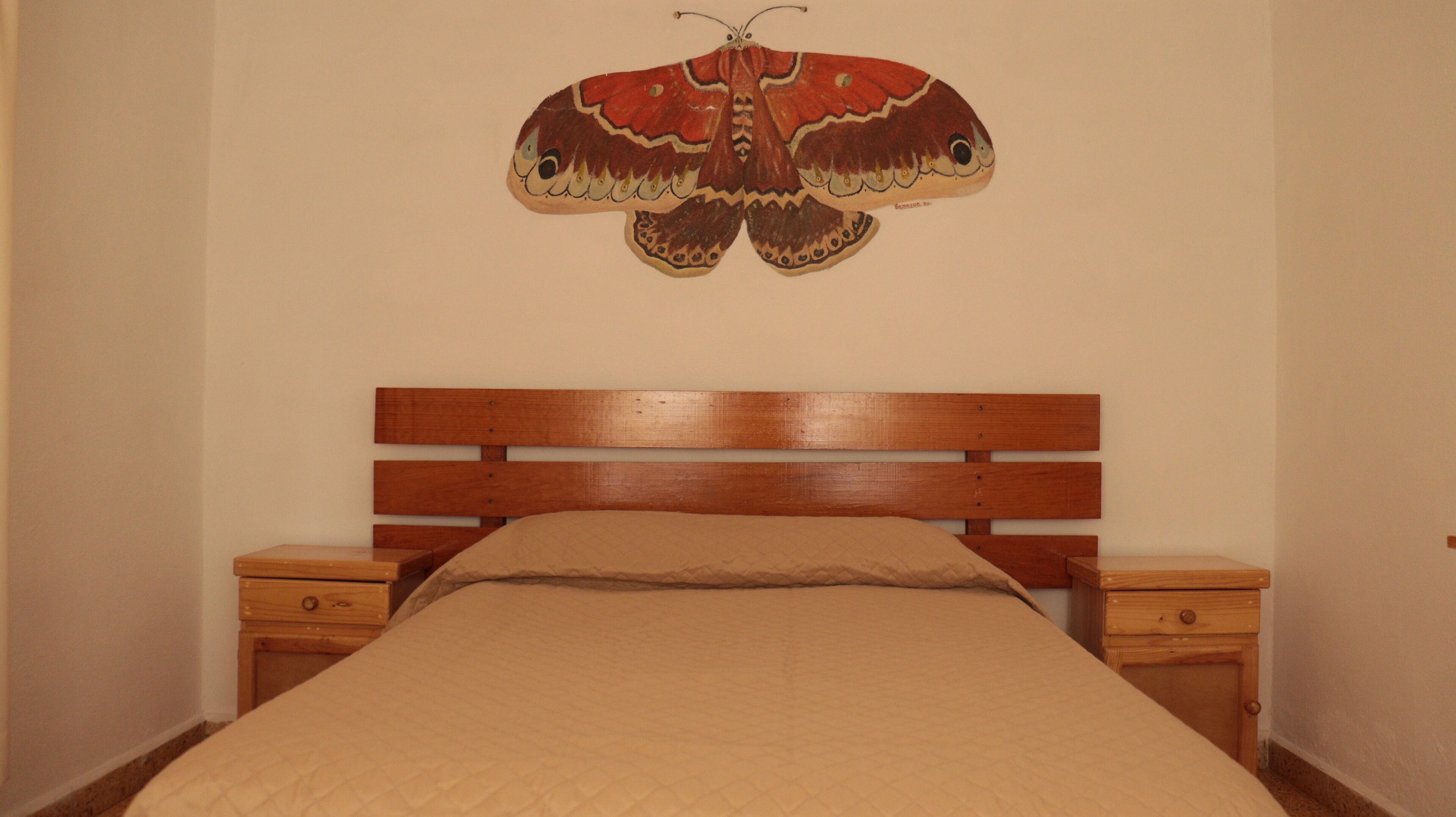 Un dormitorio con cabecera de madera, cama con colcha color beige y dos mesitas de noche con cajones.