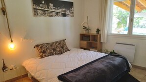Una camera da letto con un letto, un comodino, una lampada a parete, uno scaffale con oggetti decorativi e una finestra vista alberi.