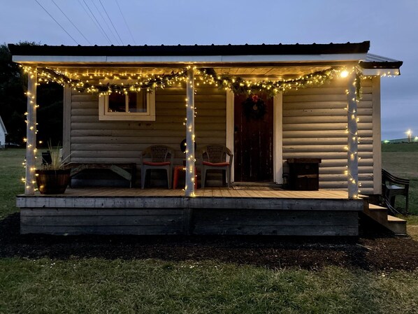 A small cabin with a porch decorated with string lights, a wooden bench, and a door with a wreath. - Bear Lodge: Romantic Tiny Cabin Near Cook Forest + Luxury Queen Bed (Marienville)