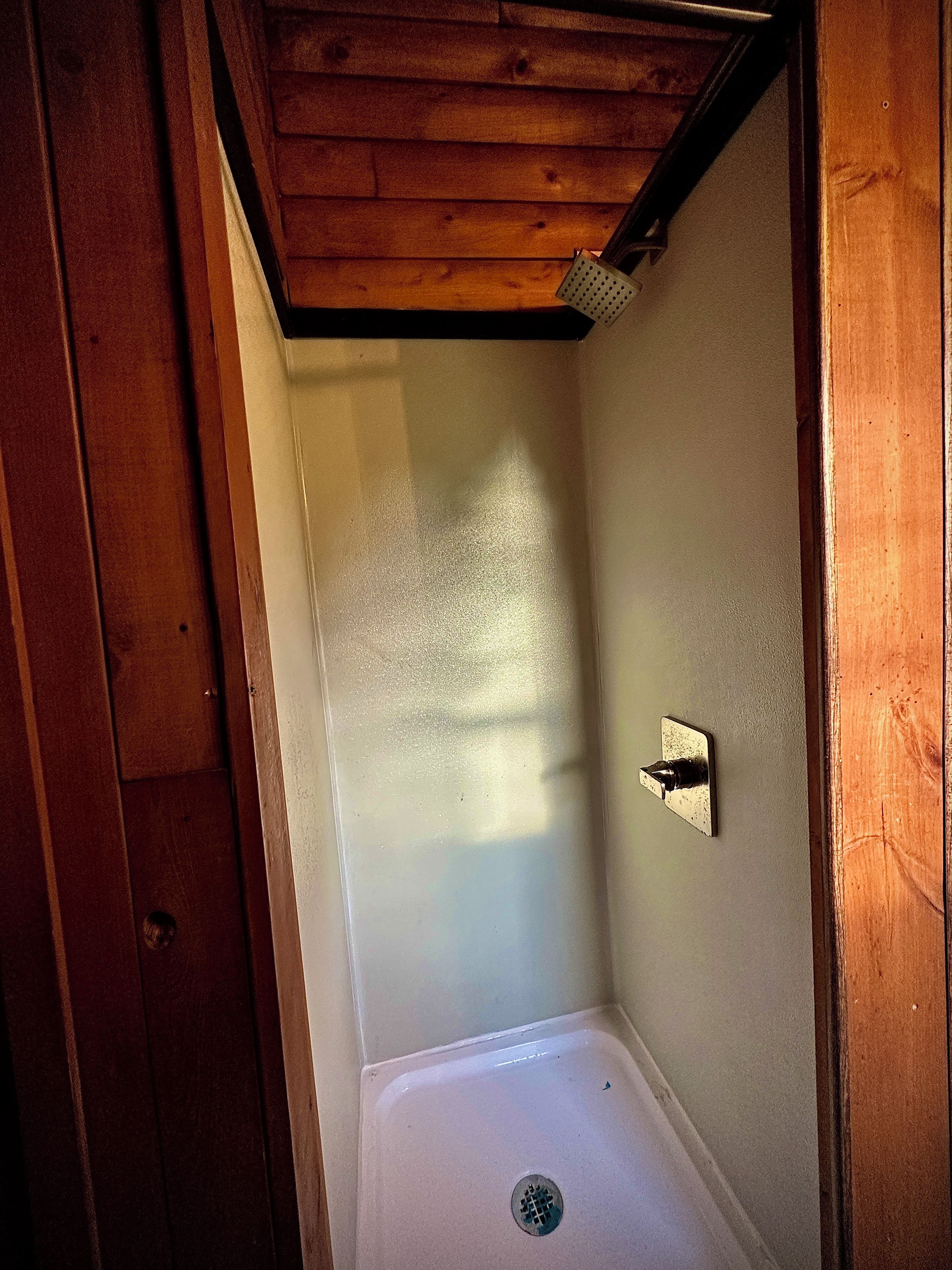 A small, enclosed shower area with a showerhead mounted on the wall, a drain in the floor, and wooden paneling on the ceiling.