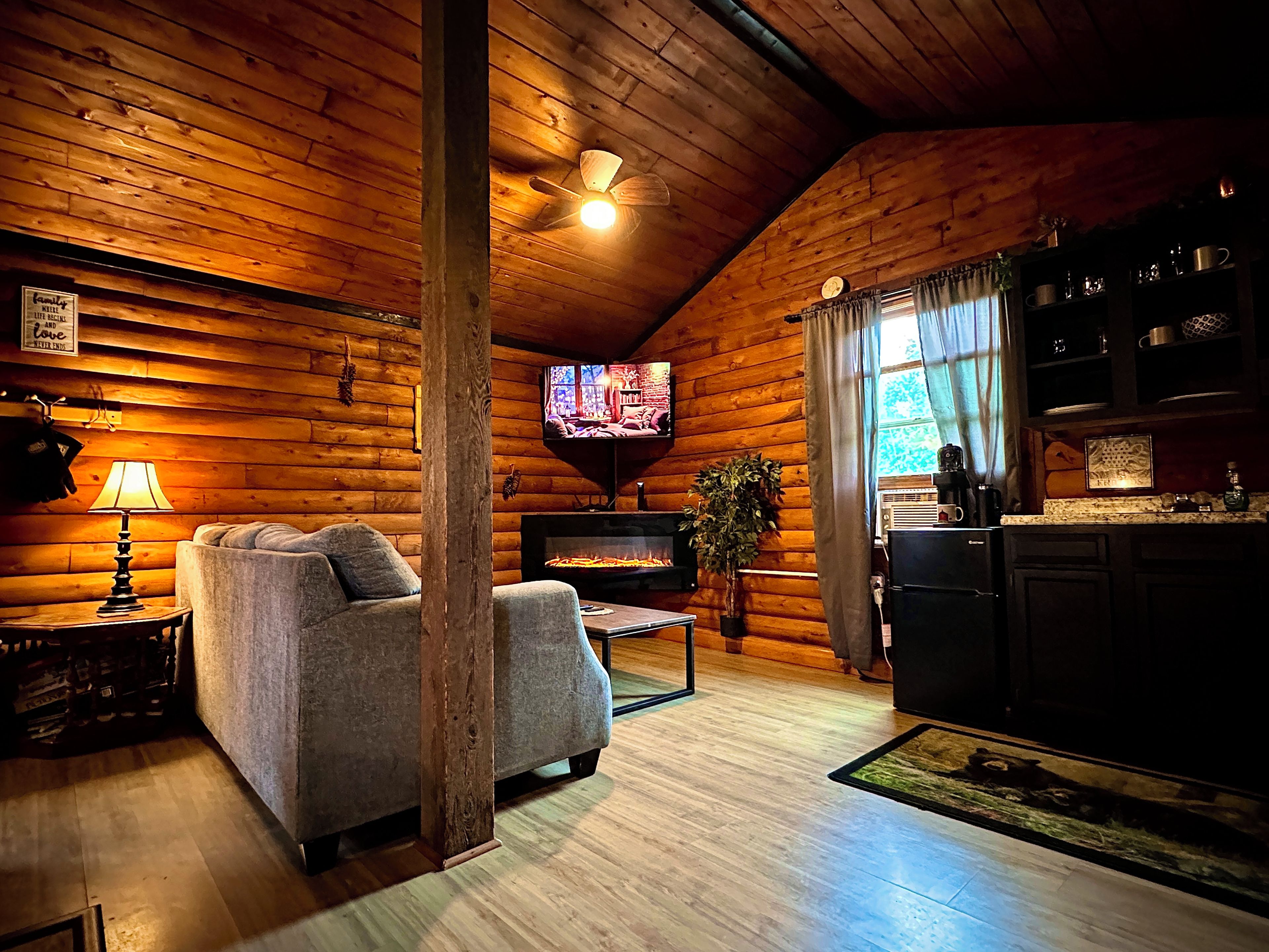 A cozy log cabin interior with a sofa, television, fireplace, and kitchen area.