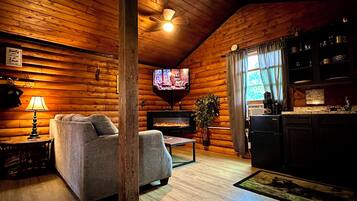 A cozy log cabin interior with a sofa, television, fireplace, and kitchen area.