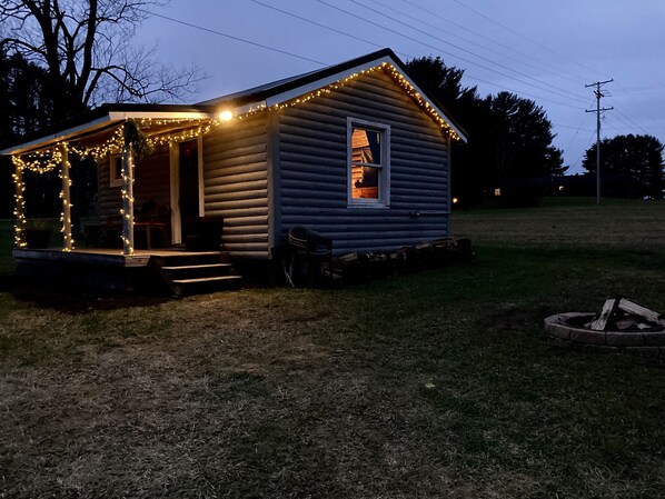 A small, single-story cabin with a porch adorned with string lights, a visible window, and a fire pit in the yard. - Bear Lodge: Pet-Friendly Tiny Cabin Near Cook Forest & Allegheny National Forest (Marienville)