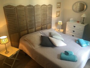 A neatly made bed with pillows and a folded towel, a bedside table with a lamp, a dresser with heart-shaped drawers, and a wall-mounted mirror.