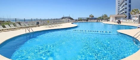 Un área de piscina con una piscina azul cristalina, un puesto de socorrista y un edificio alto al fondo.