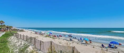 Une plage avec une clôture en bois, des parasols et des gens qui se détendent.
