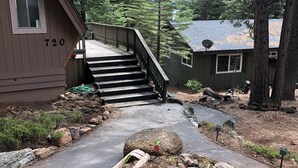 Une cabane en bois avec le numéro « 720 » peint sur le côté, un escalier menant à une terrasse, et une zone boisée avec un sentier de terre.