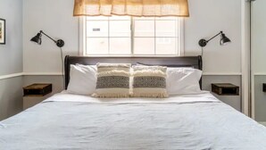 Una cama bien hecha con una colcha blanca y dos almohadas estampadas, flanqueada por dos mesitas de noche con lámparas, en una habitación con ventana y cortina.