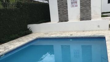 Une piscine avec un bord carrelé, un bâtiment blanc orné d’une enseigne et une haie verte en arrière-plan.