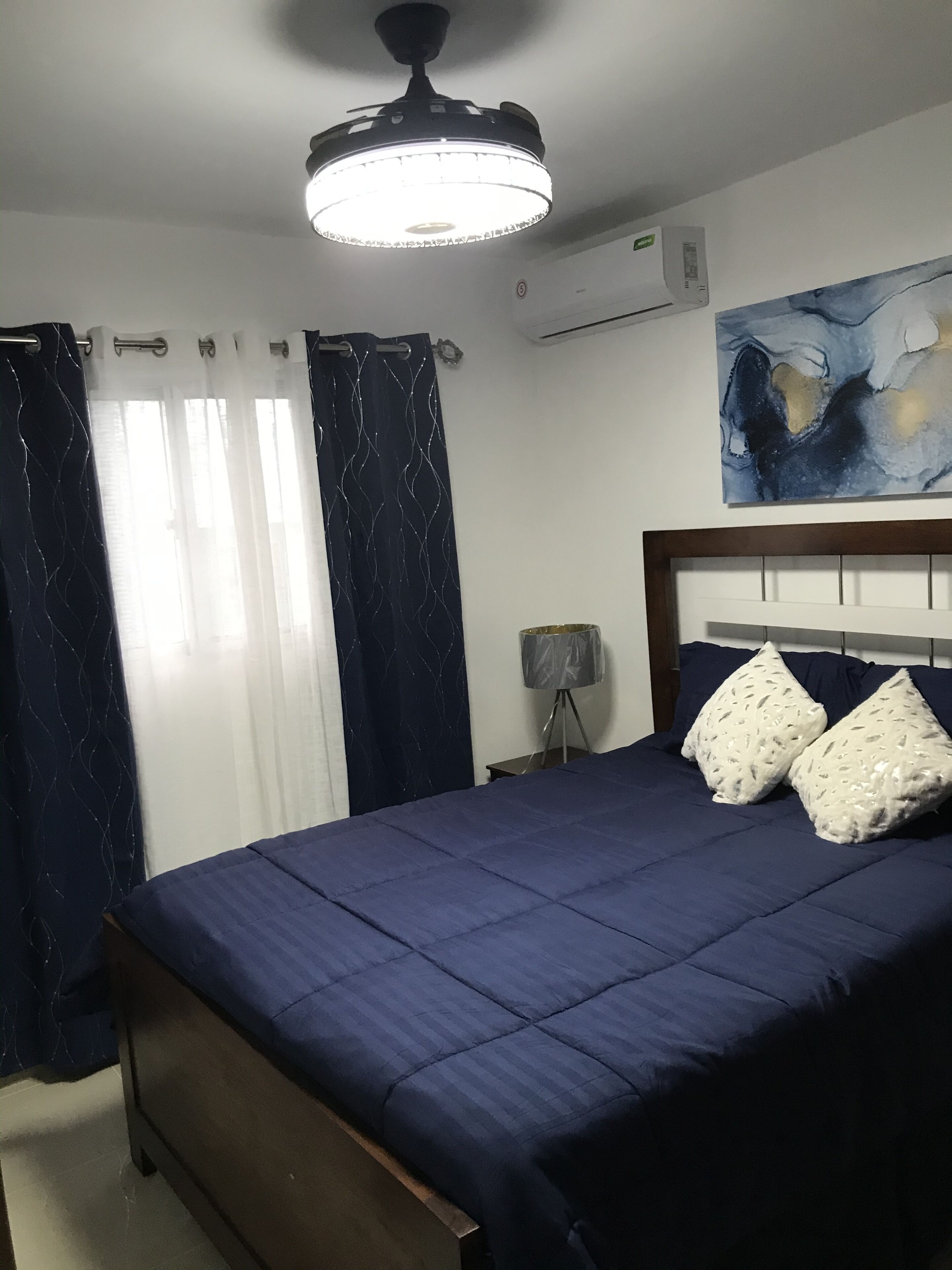 Une chambre à coucher avec un lit, des rideaux, un luminaire au plafond, un climatiseur et un tableau mural.