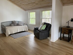 Una habitación con una cama, un sillón de cuero, una mesita de noche con una lámpara y dos ventanas.