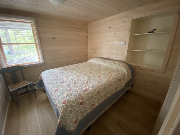 A small, cozy bedroom with a single bed, a chair, a window with blinds, and a built-in shelf.