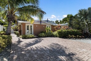 A house with a paved driveway, surrounded by lush greenery and palm trees. - Tiki Cottage: 2.5 Blocks to Beach, Pool with Swim up Tiki Bar by Anna Maria Live Vacation Rentals (Holmes Beach)