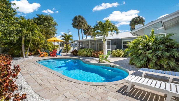 Une piscine entourée d’une terrasse en briques, de palmiers et d’une maison dotée d’un patio couvert.