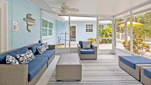 Un espace extérieur aménagé, abrité et doté de mobilier en osier, avec un canapé bleu, offrant une vue sur une piscine et une végétation tropicale.