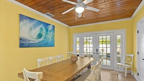 Un coin repas avec une table en bois et des chaises blanches, un ventilateur de plafond et un grand tableau représentant une vague de l’océan accroché au mur.