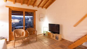 Ein gemütliches Zimmer mit großem Fenster, Korbsesseln, einem Flachbildfernseher und einer Holzbalkendecke.