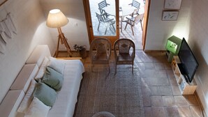 Ein gemütliches Zimmer mit einer Couch, zwei Korbstühlen, einem Fernseher und einem Fenster mit Blick auf eine Terrasse und Gartenmöbel.