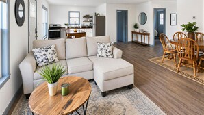 A modern living room with a sectional sofa, a dining table with chairs, a kitchen in the background, and a large mirror on the wall. - 4 Bedroom Lakehouse w/ Hot Tub, Firepit, & Game Room (Cadiz)