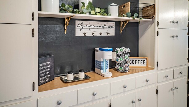 Une petite cuisine avec des meubles blancs, un coin café et une pancarte sur laquelle on peut lire : « My cupunnethover ».