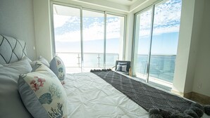 Ett sovrum med en stor säng, utsikt över havet genom fönster som sträcker sig från golv till tak och en balkong.