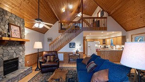 Ein geräumiges Wohnzimmer mit Kamin aus Stein, einem Ledersessel, einem blauen Sofa und einer Holztreppe, die zu einer oberen Ebene führt.