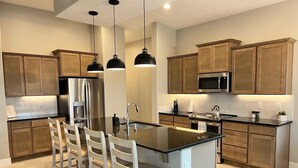 A modern kitchen with wooden cabinets, a stainless steel refrigerator, a microwave, and a large island with bar stools. - Chill. Grill. Putt. Splash. Play. Your Private Pool Paradise Awaits! (Mesquite)