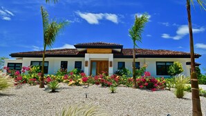 A modern house with a tiled roof, surrounded by a gravel driveway and colorful flowering plants. - Casa Palmeras • Luxury Villa Near Beach & Surf (Tamarindo)