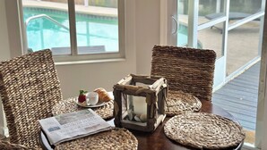 A wicker chair with a newspaper, a candle, and a croissant on a wooden table by a window overlooking a pool. - Serenity Waterfront 4BR w/ Private Dock, Heated Pool & Relaxing Hot Tub (Palm Coast)