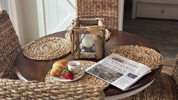 A round wooden table with woven placemats, a newspaper, a croissant, a cup of coffee, and a rustic lantern. - Serenity Waterfront 4BR w/ Private Dock, Heated Pool & Relaxing Hot Tub (Palm Coast)