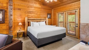 A wooden cabin bedroom with a bed, a nightstand, a lamp, a ceiling fan, and a door with a view of the outside. - Romantic Studio Cabin for Two with Private Balcony, Jetted Tub, and Gas Fireplace (Branson)