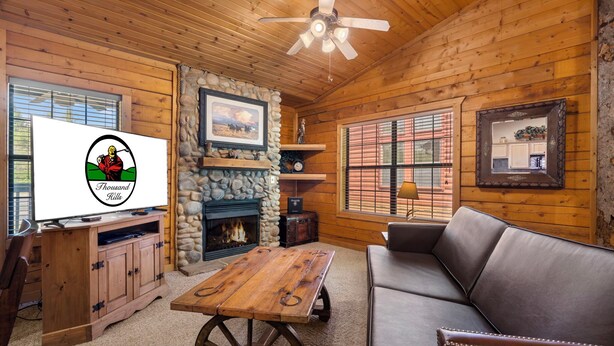 A cozy cabin interior with a stone fireplace, wooden furniture, and a television displaying a logo. - Romantic Studio Cabin for Two with Private Balcony, Jetted Tub, and Gas Fireplace (Branson)