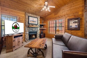 Ein gemütliches Blockhaus-Interieur mit Kamin aus Stein, Holzmöbeln und einem Fernseher, der ein Logo zeigt.