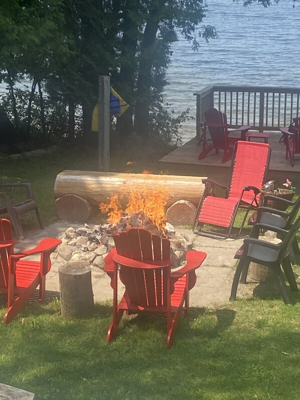 An outdoor seating area with a fire pit surrounded by red chairs, a wooden deck, and a view of a lake.