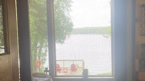 A view through a window with a metal screen, a wooden door frame, and a view of a red chair and table outside. - Tranquil 3 bedroom waterfront cottage on private lake! (Rideau Lakes)