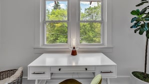 A white desk with two drawers and a candle on it, positioned in front of a window with a view of trees.