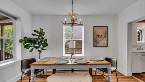 A dining area with a wooden table, chairs, a chandelier, a plant, and a window with a view of trees.